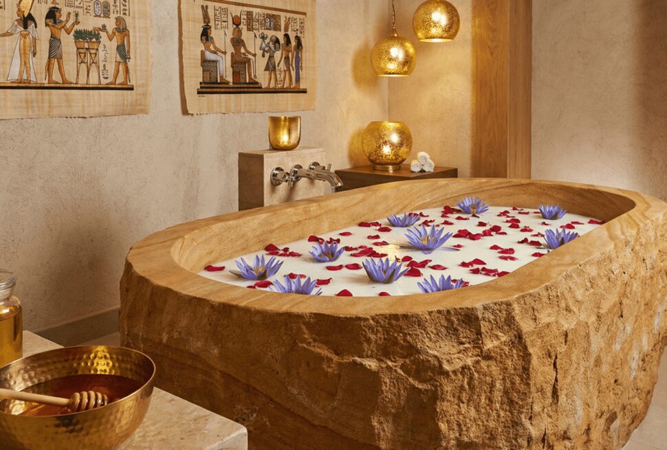 A luxurious stone bathtub filled with milk, rose petals, and lotus flowers, representing the ancient Egyptian Cleopatra beauty ritual in a spa setting.